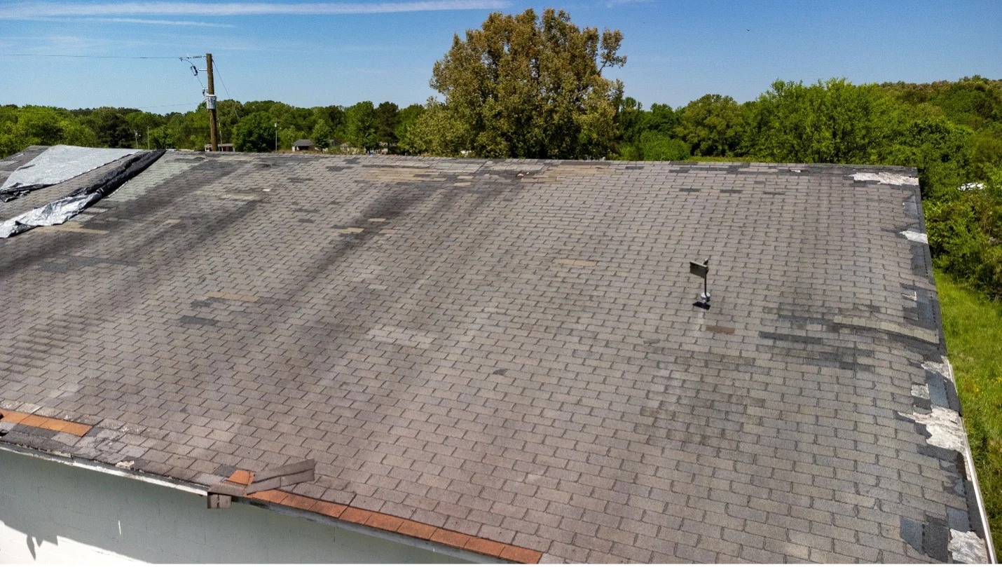 A large roof with gray asphalt shingles, showing visible wear, missing shingles, and patches of damage. Some shingles are discolored or curling. Trees and utility poles are visible in the background under a clear sky.
