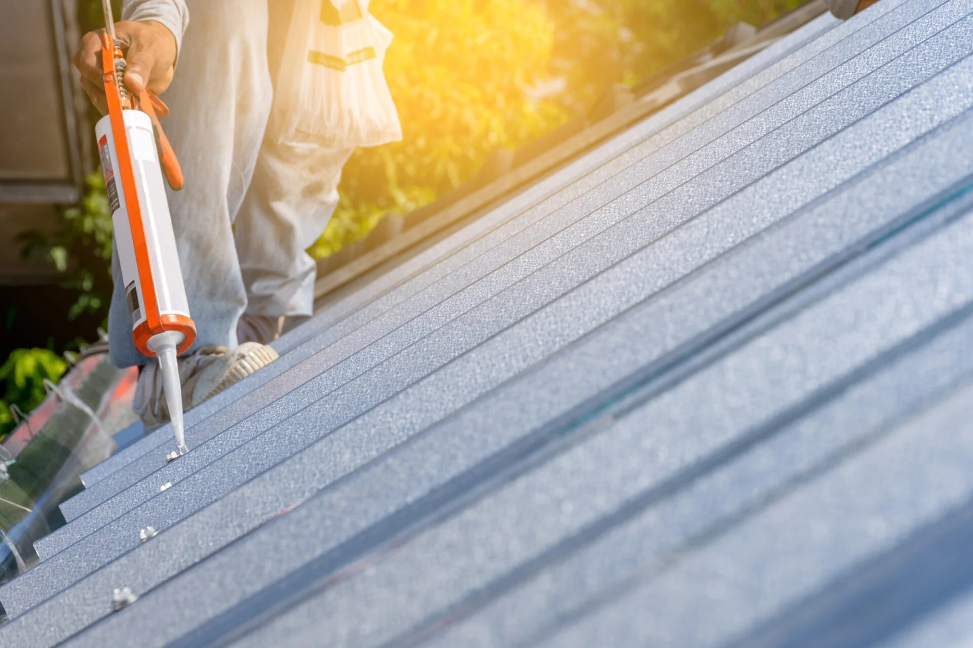 A person applies metal roof sealant to the joints of a metal roof with a caulking gun on a sunny day, ensuring roof protection as sunlight shines on the panels.