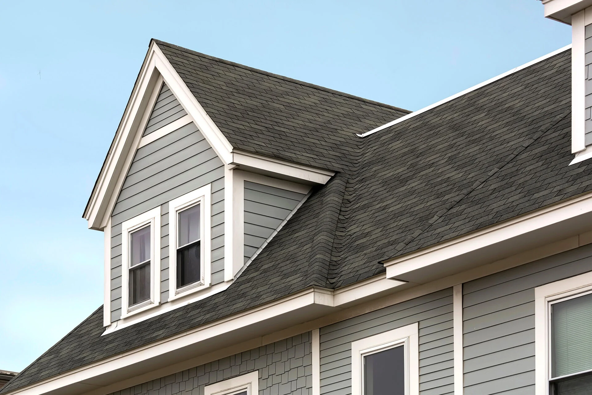 A close-up view of a house with gray siding, white trim, and a steeply pitched roof featuring asphalt shingles and double-hung windows under a clear blue sky.