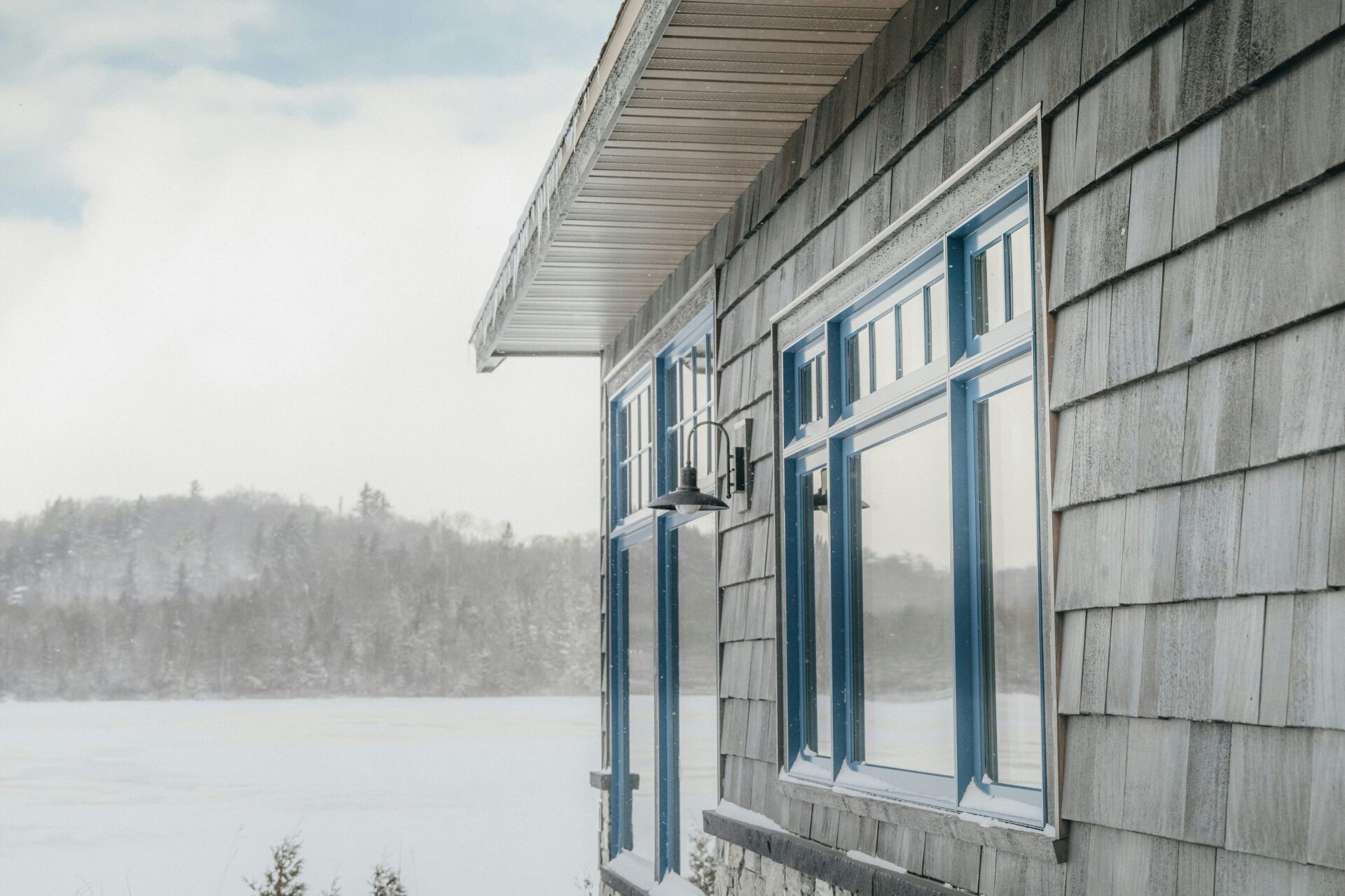 A house with blue window frames by a frozen lake. Snow covers the ground and trees in the background.