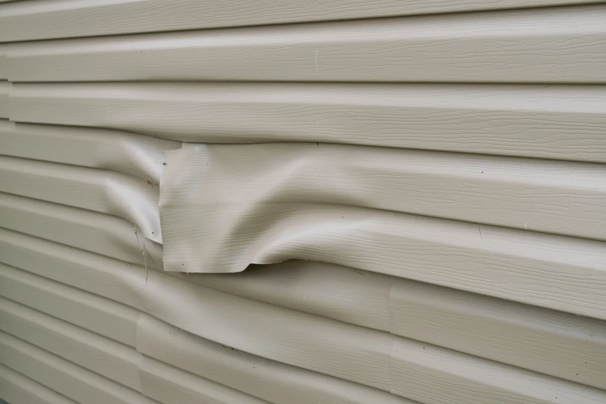 Close-up of beige vinyl siding with visible dents and warping, showing significant damage to several horizontal panels and highlighting the need for vinyl siding repair.