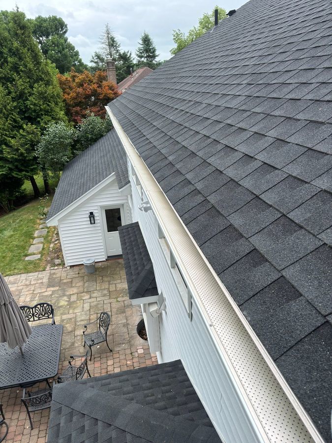 View from a rooftop showing clean black shingles, white gutters, and a patio below with a metal table and chairs, surrounded by trees and greenery.