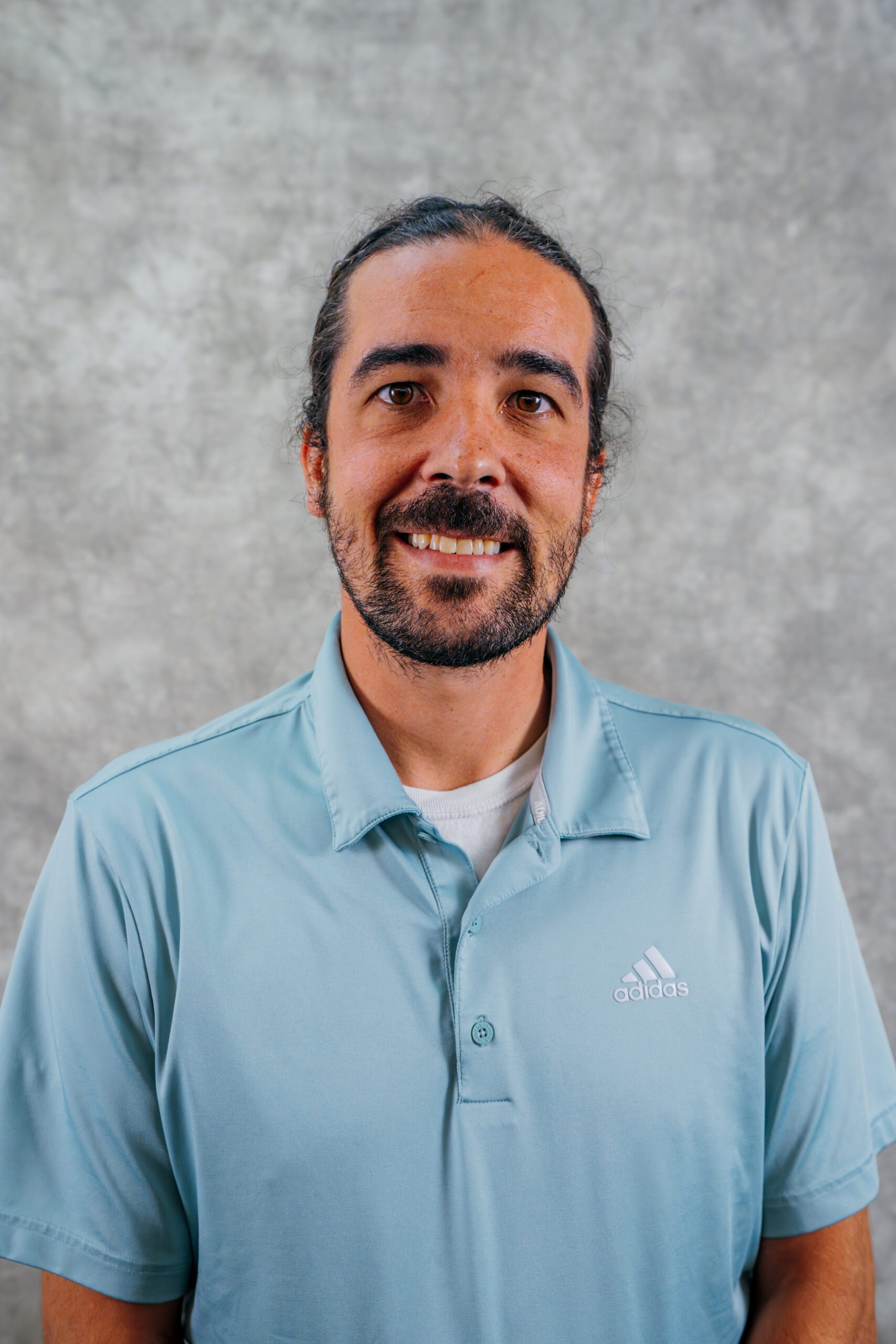 Eric Houchin, a man with long dark hair tied back, a beard, and mustache, is smiling. He is wearing a light blue Adidas polo shirt and standing in front of a mottled gray background.