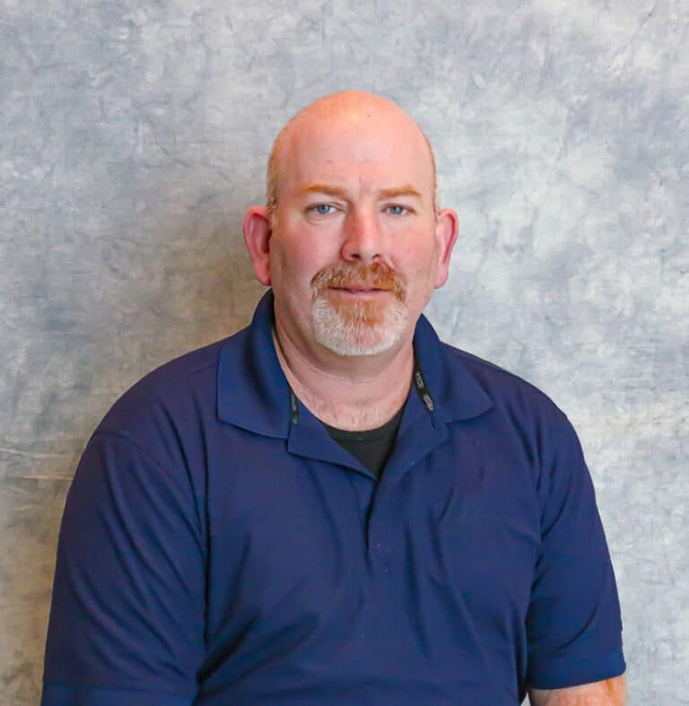 Bald Man with Moustache in Blue Polo Shirt A bald man with a moustache wearing a blue shirt. He is standing against a grey background.