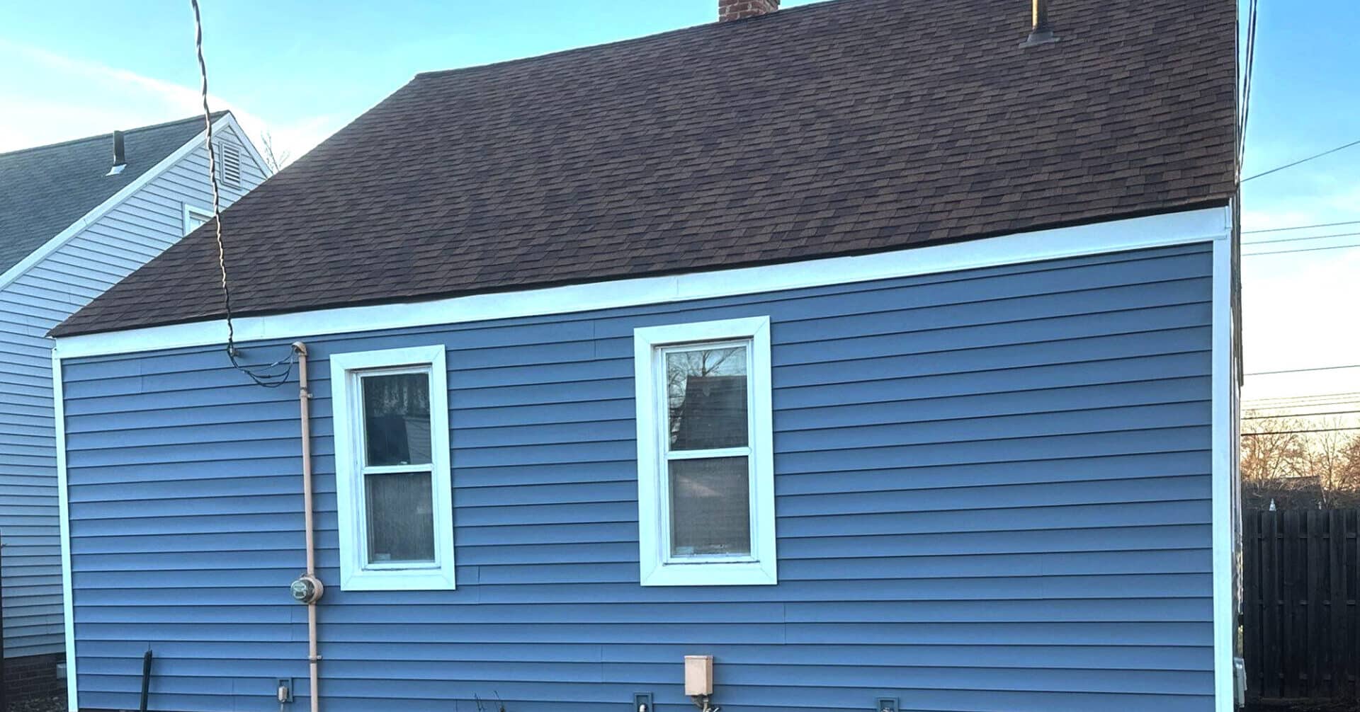 A blue house with two windows and brown roof. The house has white trim around the windows and roof.