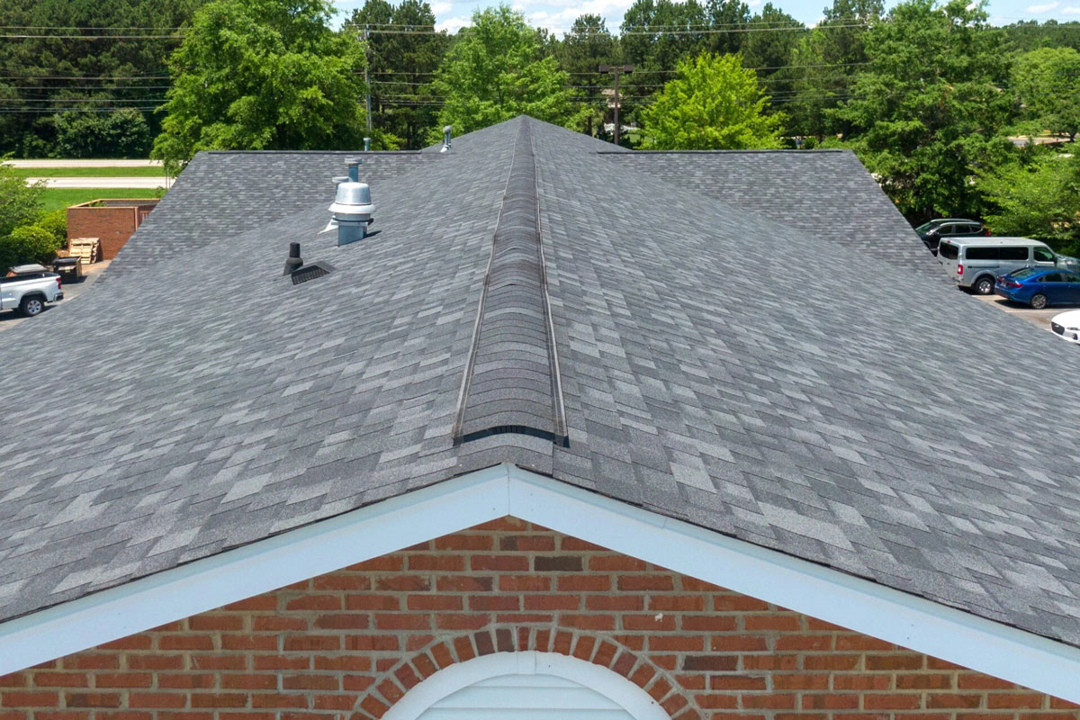 Roof of a brick building with grey shingles A grey roof with a brick building in the foreground. The roof has a small vent on it.