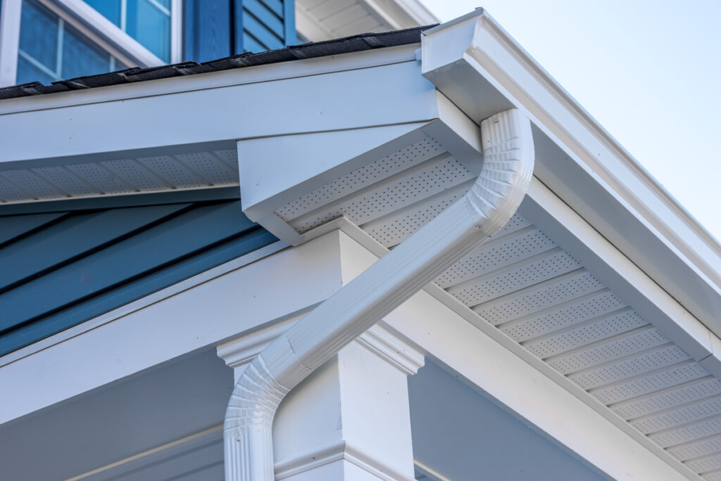 Close-up of a house corner showing a white gutter and downspout attached to the roof, with blue siding and white trim. The soffit features small vent holes for attic ventilation.