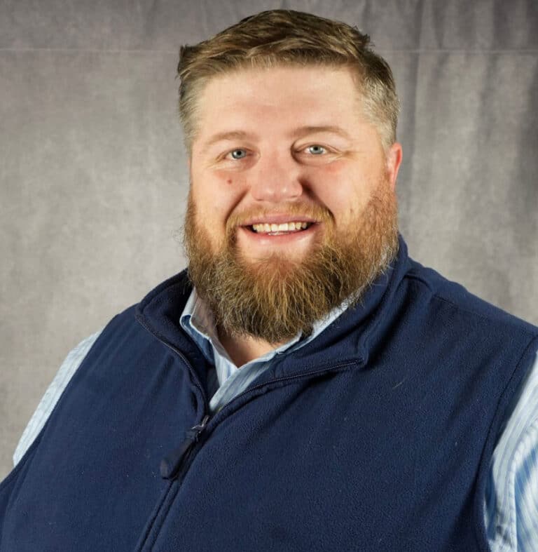 Smiling man with beard and blue vest A man with a beard smiles at the camera. He is wearing a blue vest.