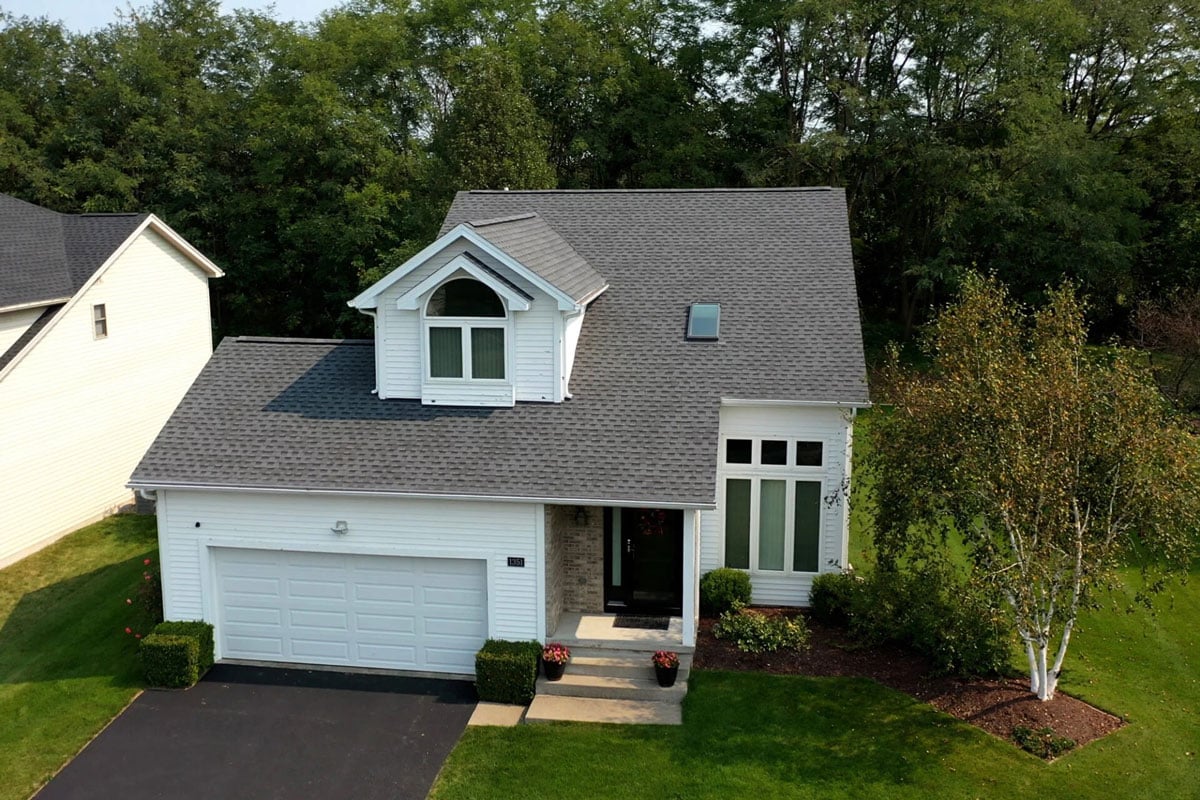 Aerial view of a house with a gray roof and white garage door. The house has a well-manicured lawn and trees.