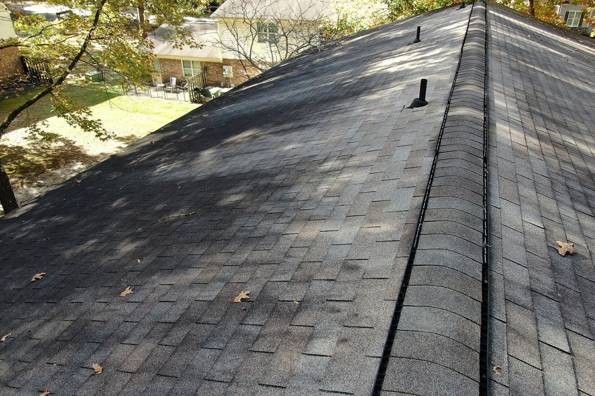 Roof with black shingles. A house and trees are visible in the background.