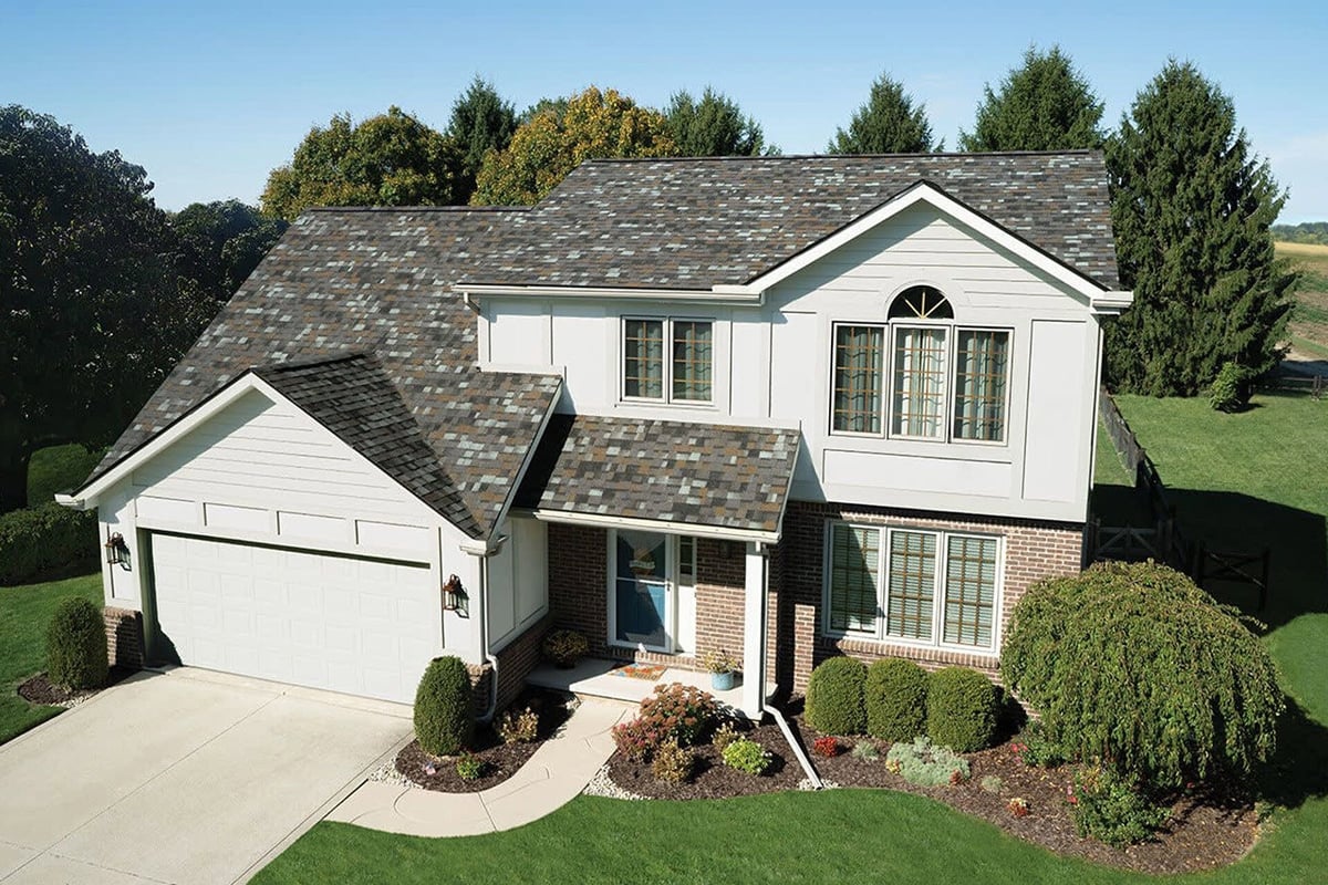 Aerial view of a two-story house with a gray roof and white siding. The house has a well-manicured lawn and shrubs.
