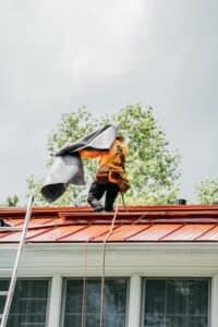 A worker in orange safety gear installing a roof vent. The worker is on a ladder.