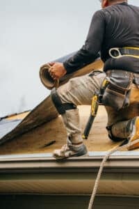 A roofer in a tool belt working on a roof. The worker is focused on the task.