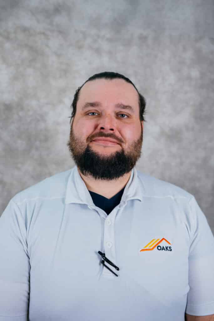 A man with a beard wearing a white polo shirt. He is standing in front of a gray background.