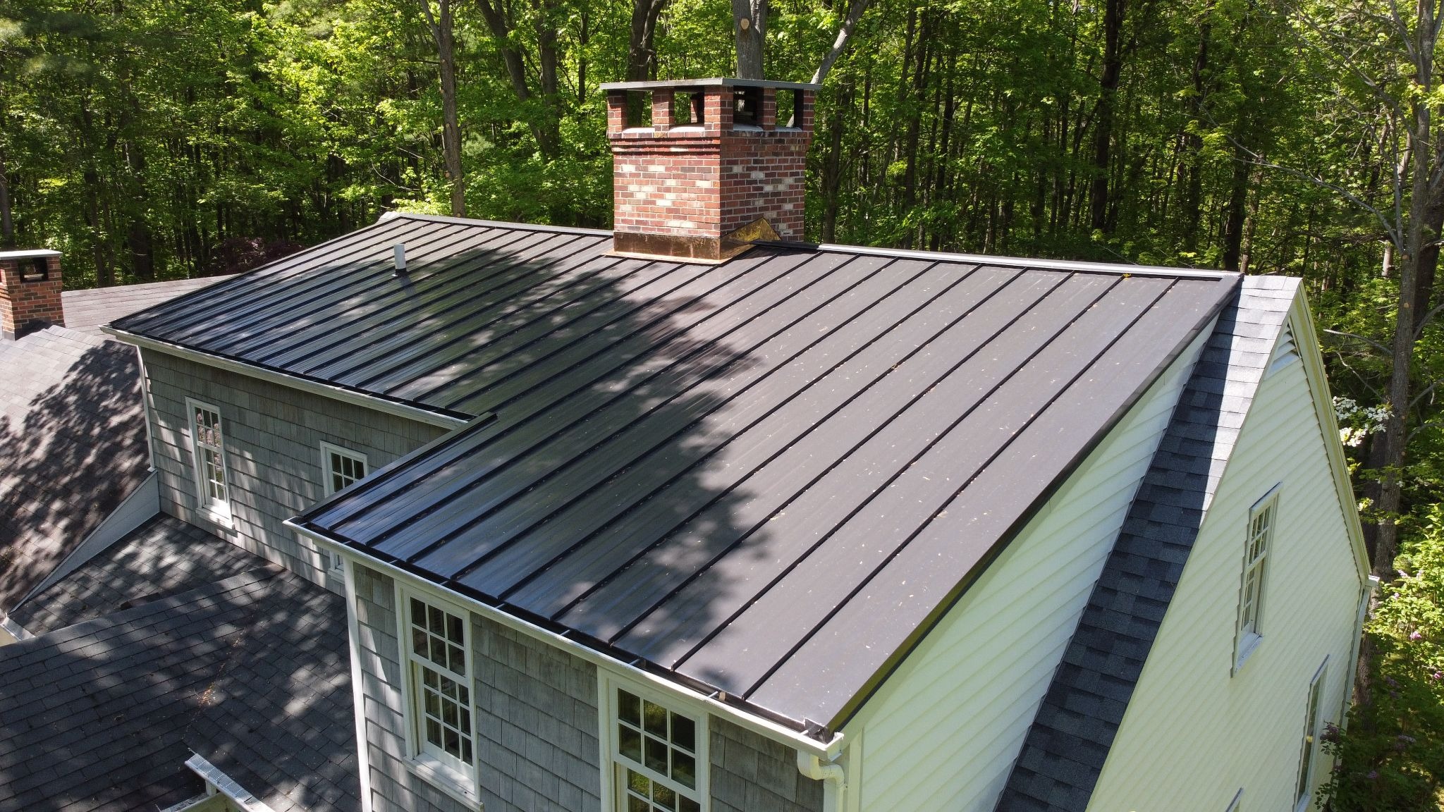 A house with a metal roof and a brick chimney surrounded by trees. The house has white and gray siding.