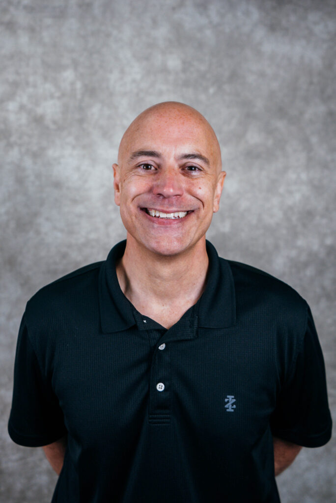 A bald man smiling and wearing a black polo shirt. He appears to be in a studio setting.