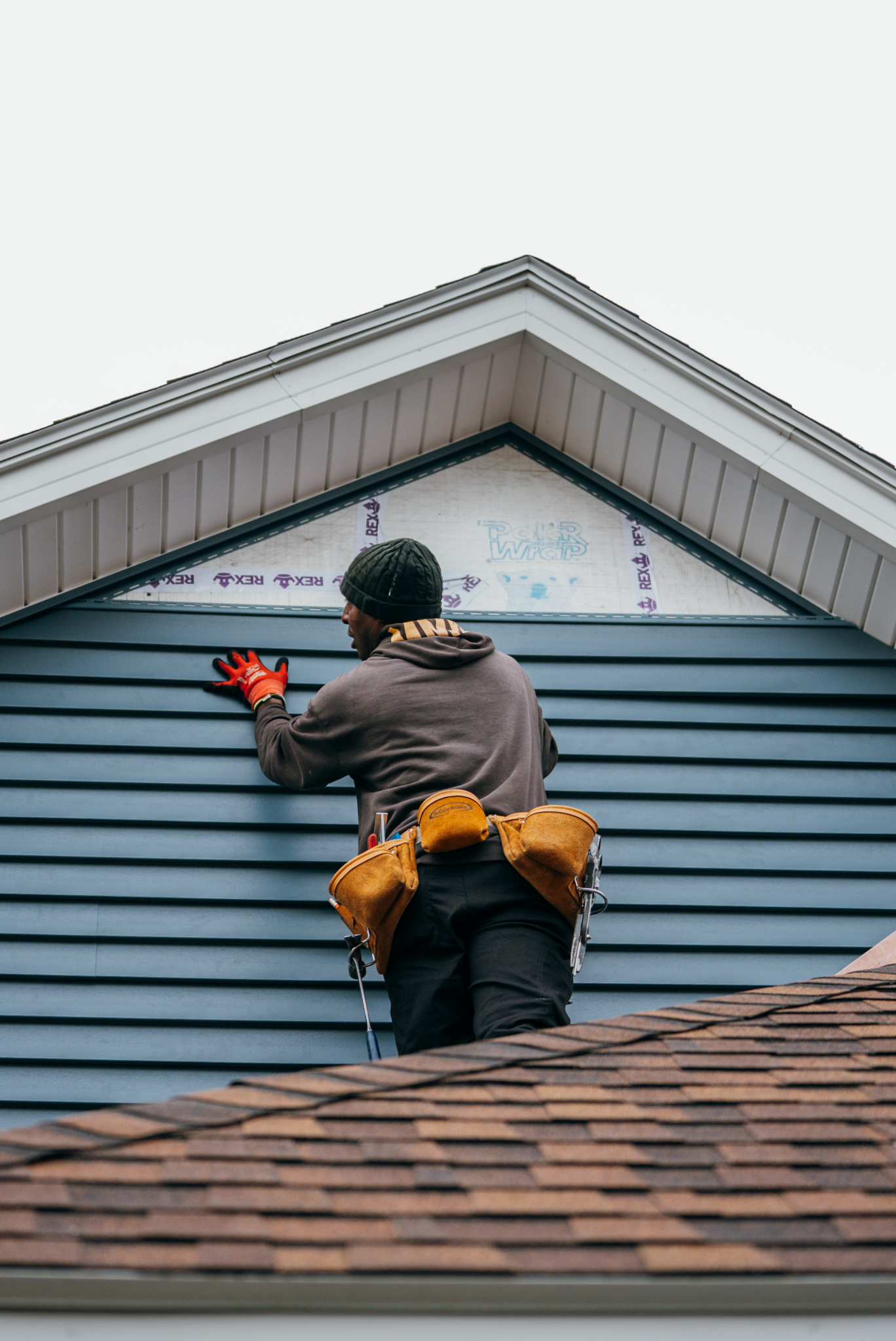 A worker in a brown jacket installing siding on a roof. The worker is wearing a tool belt.
