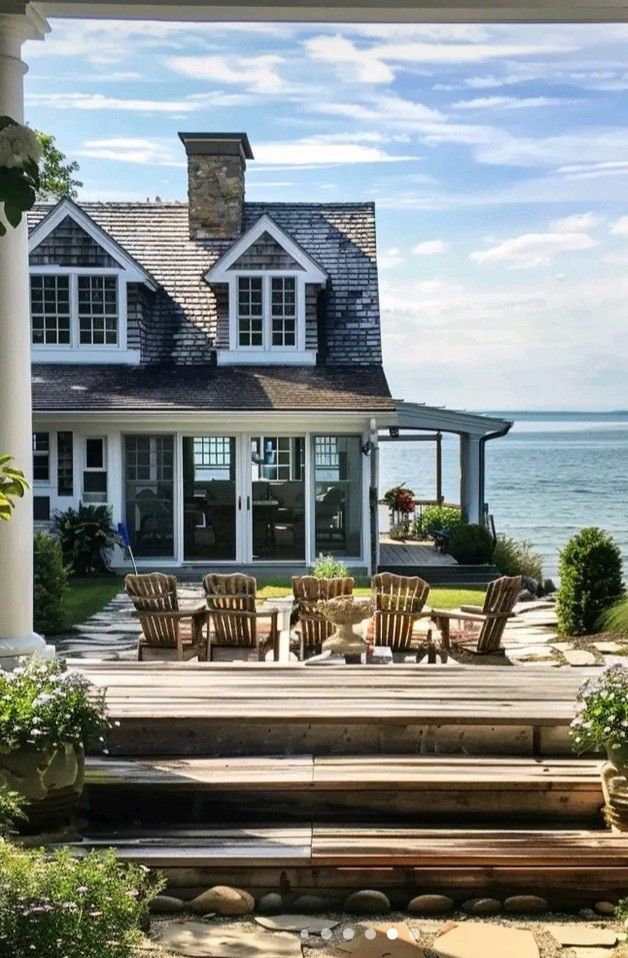 A large house with a deck overlooking the ocean. The deck has wooden chairs and a table.