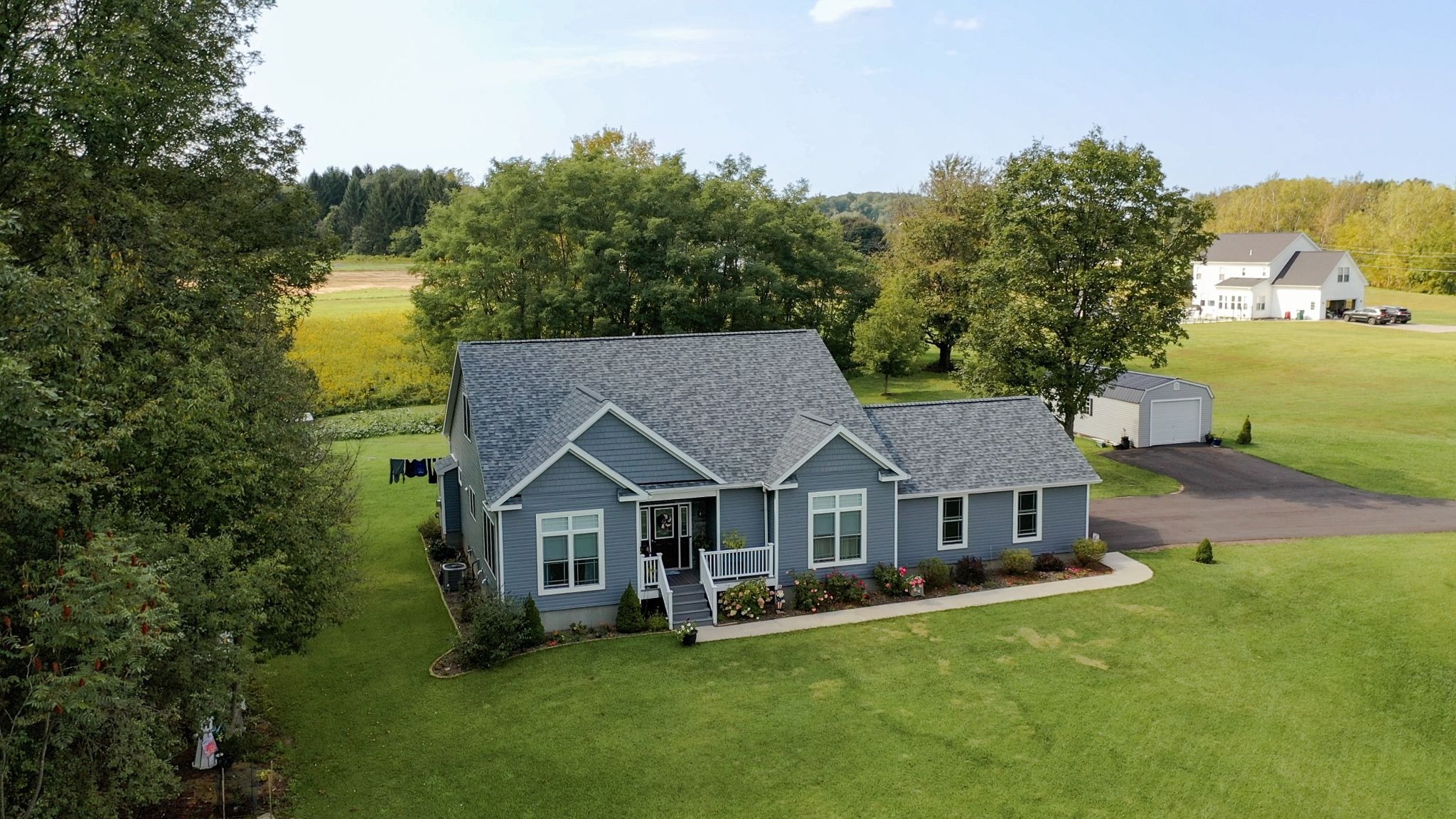A single story blue house with a gray roof sits on a large green lawn with trees and a driveway leading to a white garage.
