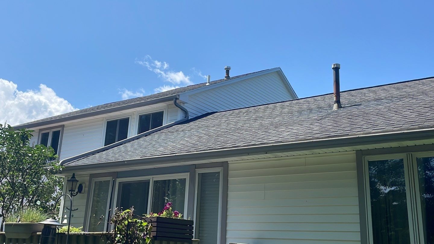 The roof of a house with white siding and a gray shingle roof. A two-story section of the house is visible above the roof.