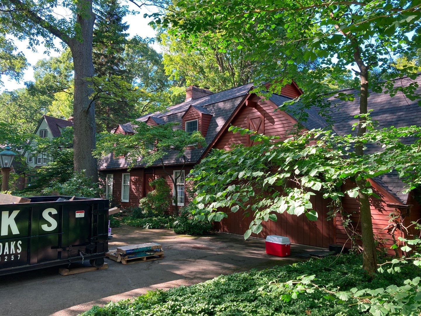 A large red house with a dumpster in the driveway surrounded by trees and greenery. The house has a brown roof and multiple windows.