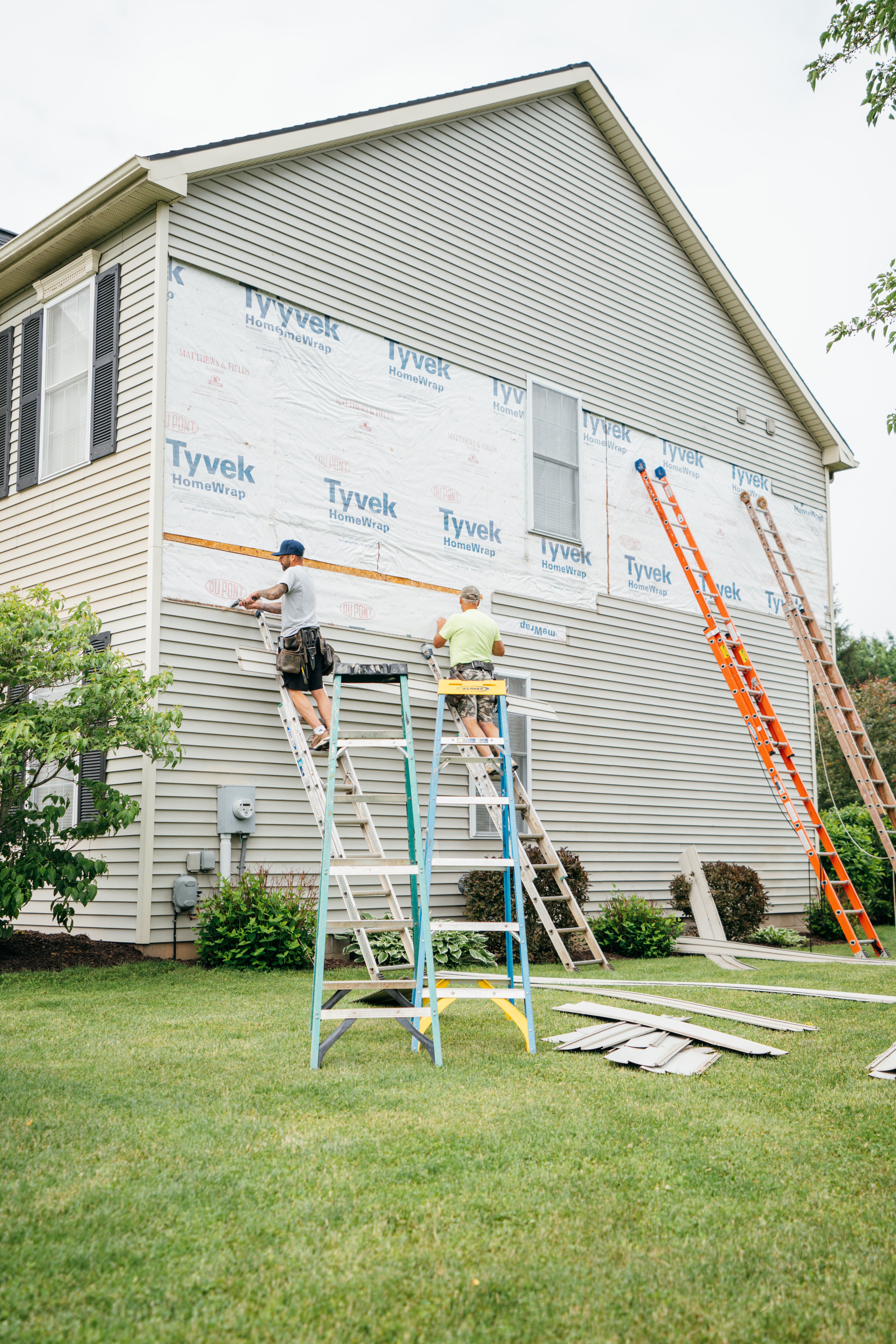Two workers on ladders installing exterior siding on a house with Tyvek wrapping.
