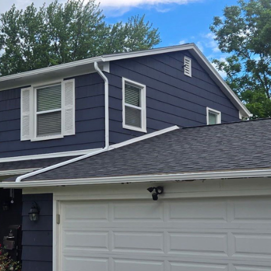 A two-story house featuring blue siding, white window shutters and trim, a grey shingle roof, and a white garage door surrounded by green trees.