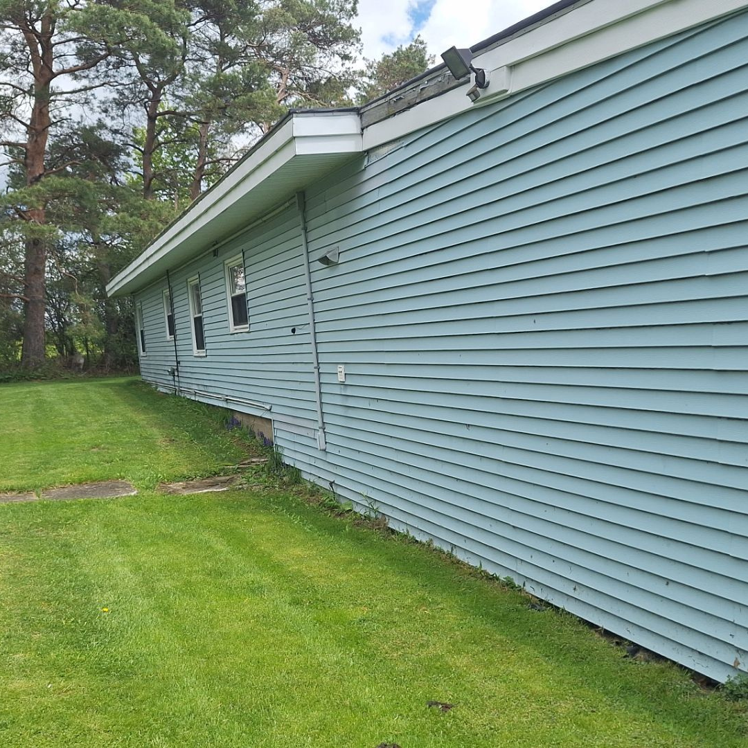A light blue house with horizontal siding features a green lawn and tall trees in the background.