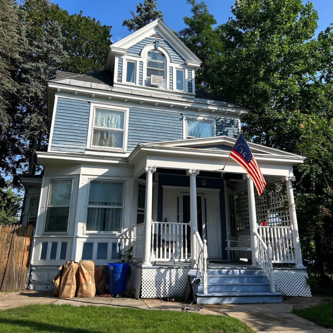 Charming blue Victorian-style home featuring a white porch, American flag, and front yard with green lawn. The house has decorative siding, dormer windows, and lattice accents.