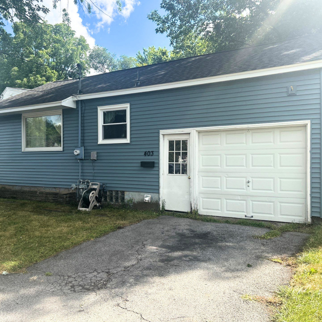 A blue garage with a white door and adjacent white entrance. The driveway leads up to the garage on a sunny day.