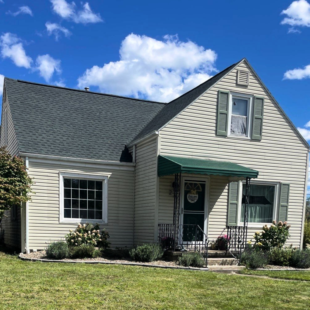 A cozy beige house with a green awning and shutters, surrounded by well-maintained greenery and a clear blue sky.