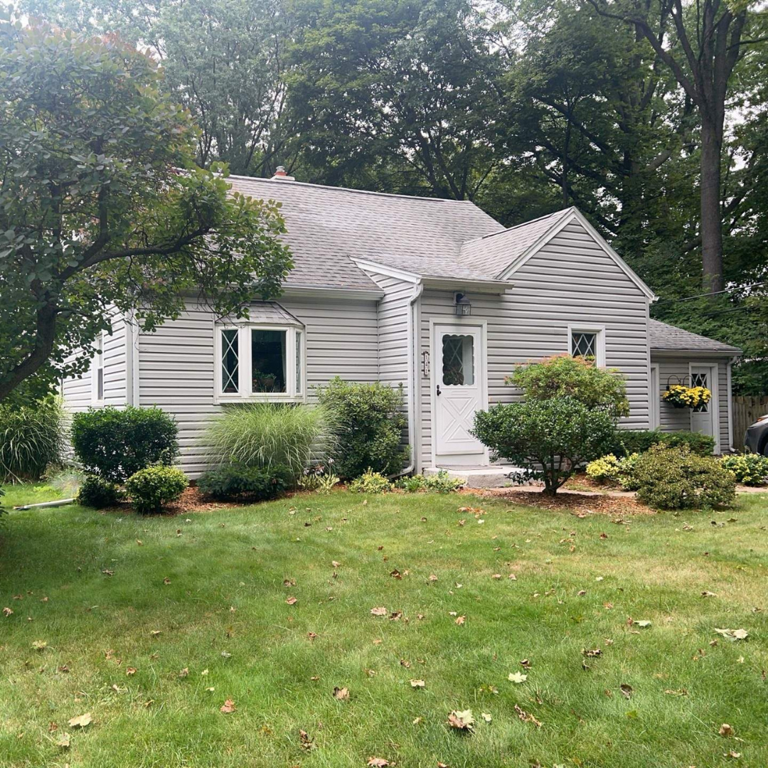 A gray house with white trim surrounded by well-maintained green lawn, shrubs, and trees.