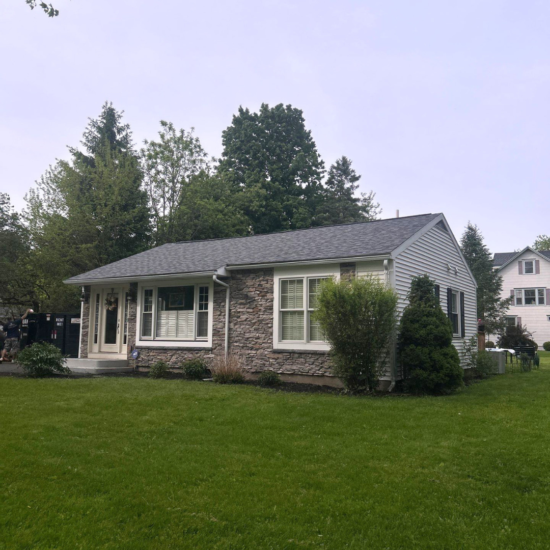 A cozy single-story house featuring a mix of stone and white siding, surrounded by lush greenery. The front yard is well-maintained with a neatly trimmed lawn and decorative shrubs.