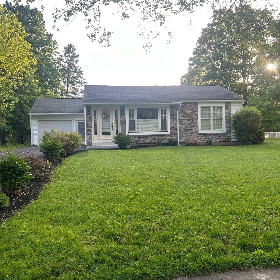 A charming single-story house with a stone facade, white trim, and a well-maintained green lawn surrounded by trees. The front yard features neatly trimmed bushes and a driveway leading to a garage on the left side.