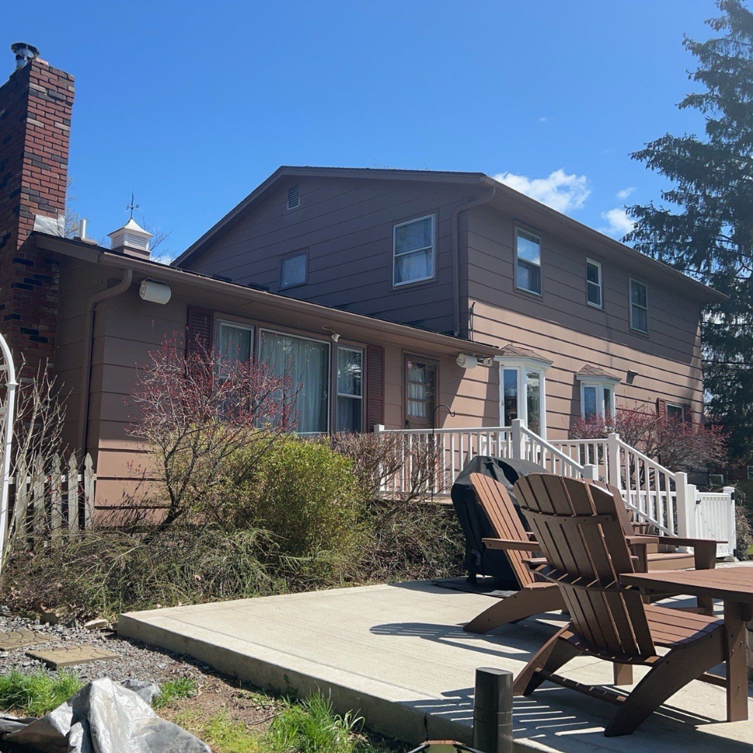 Charming backyard featuring wooden patio chairs on a small deck, with a house in the background. The scene is bright and sunny with greenery around.