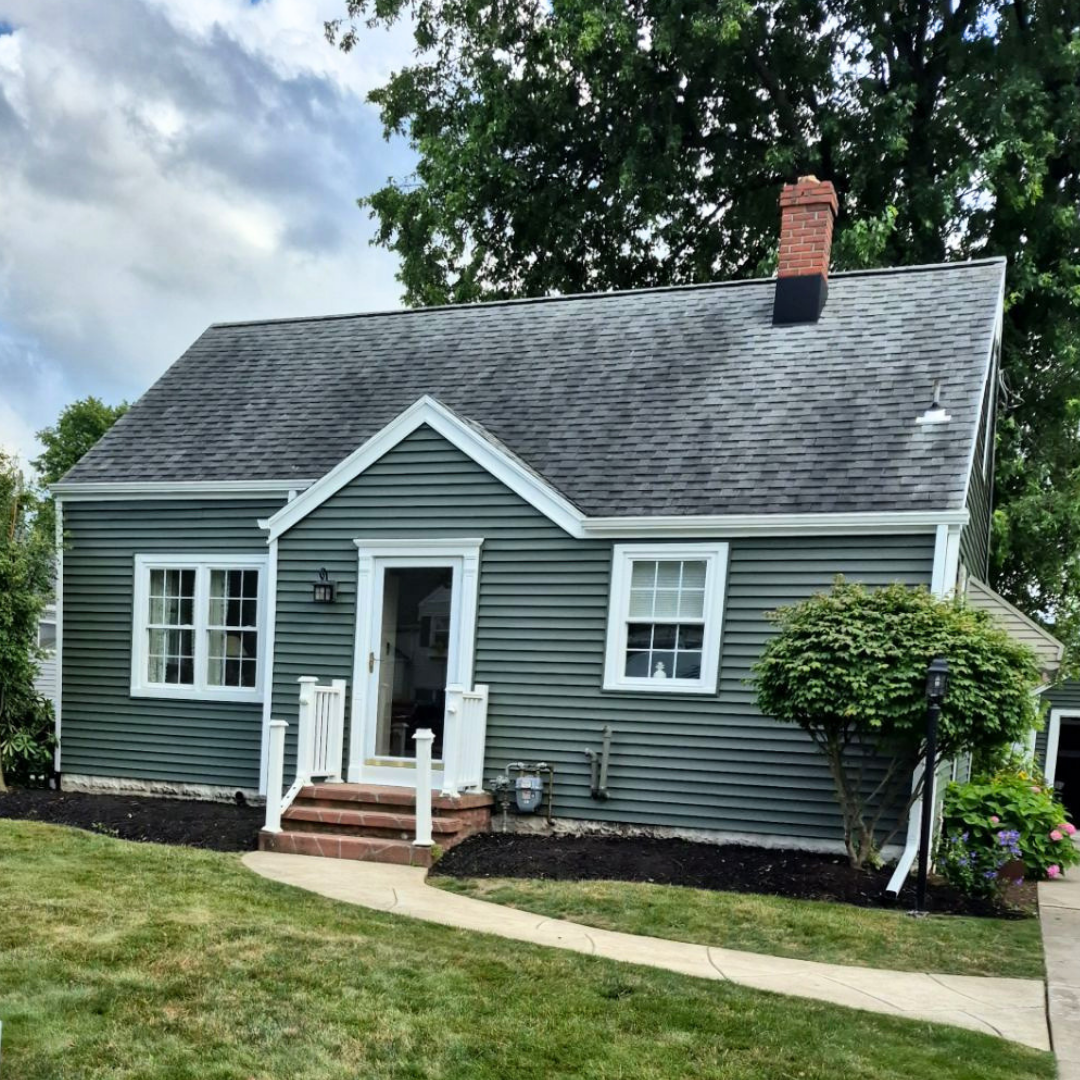 A quaint green house featuring a newly installed gray roof, white trim, and a welcoming brick pathway leading to the front door, surrounded by lush greenery under a partly cloudy sky.