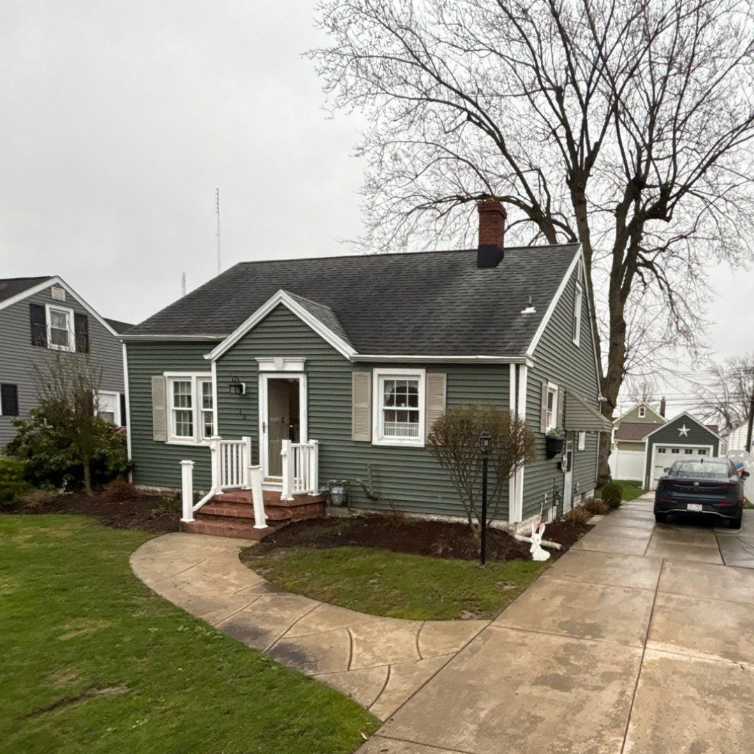 A green house featuring a curved concrete walkway leading to the front door, surrounded by a neat lawn. A black car is parked in the driveway, and leafless trees add a rustic touch.