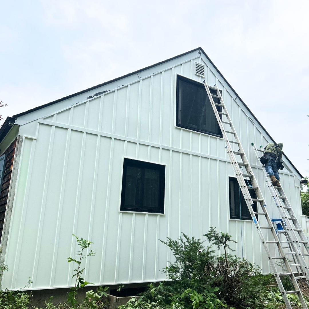 A worker climbs a ladder to install white house siding on a two-story structure with black windows. The scene highlights the exterior renovation process and craftsmanship involved.