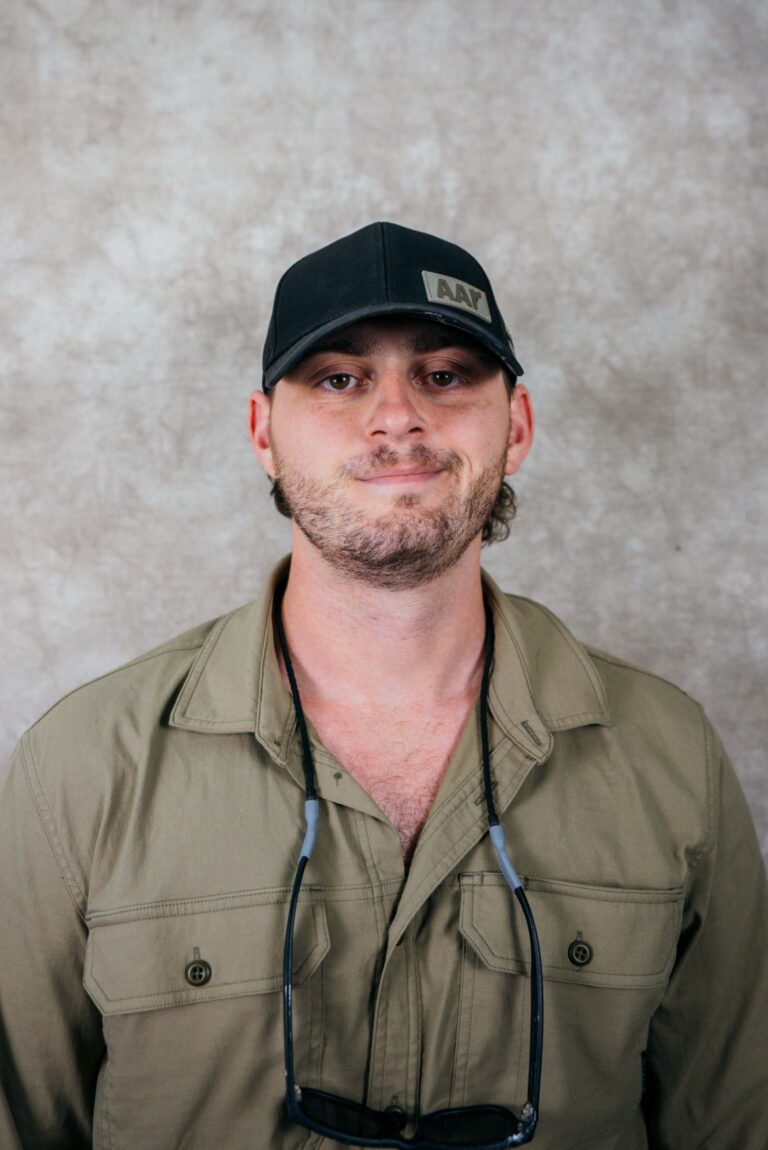 Portrait of a man in a black cap and khaki shirt. He has neck-mounted glasses and stands against a neutral background.