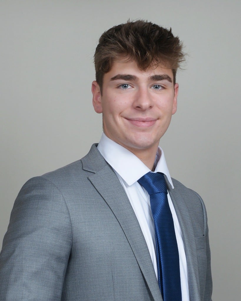 Man in a gray suit with a white shirt and blue tie smiling at the camera; close-up image of his attire.