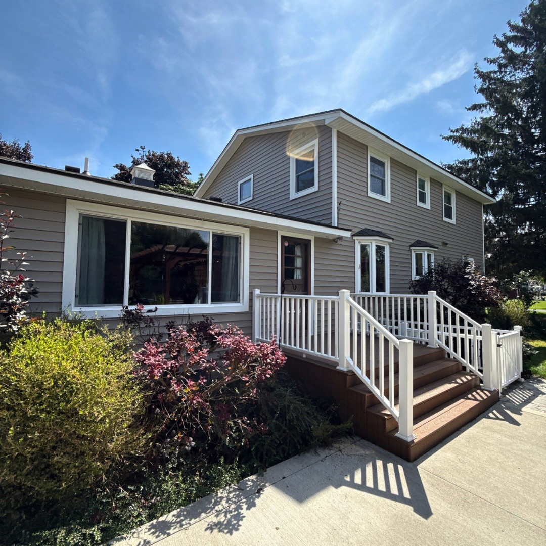 Exterior view of a modern home featuring a white deck and wooden stairs leading to the entrance, surrounded by greenery.