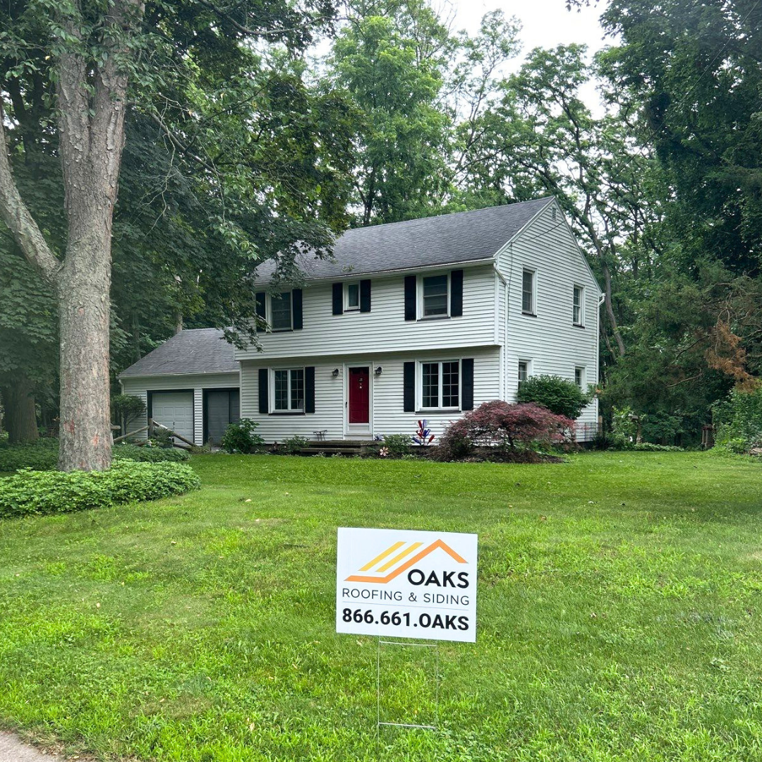 White house with Oaks Roofing & Siding yard sign. Green surroundings and a tree in the foreground.