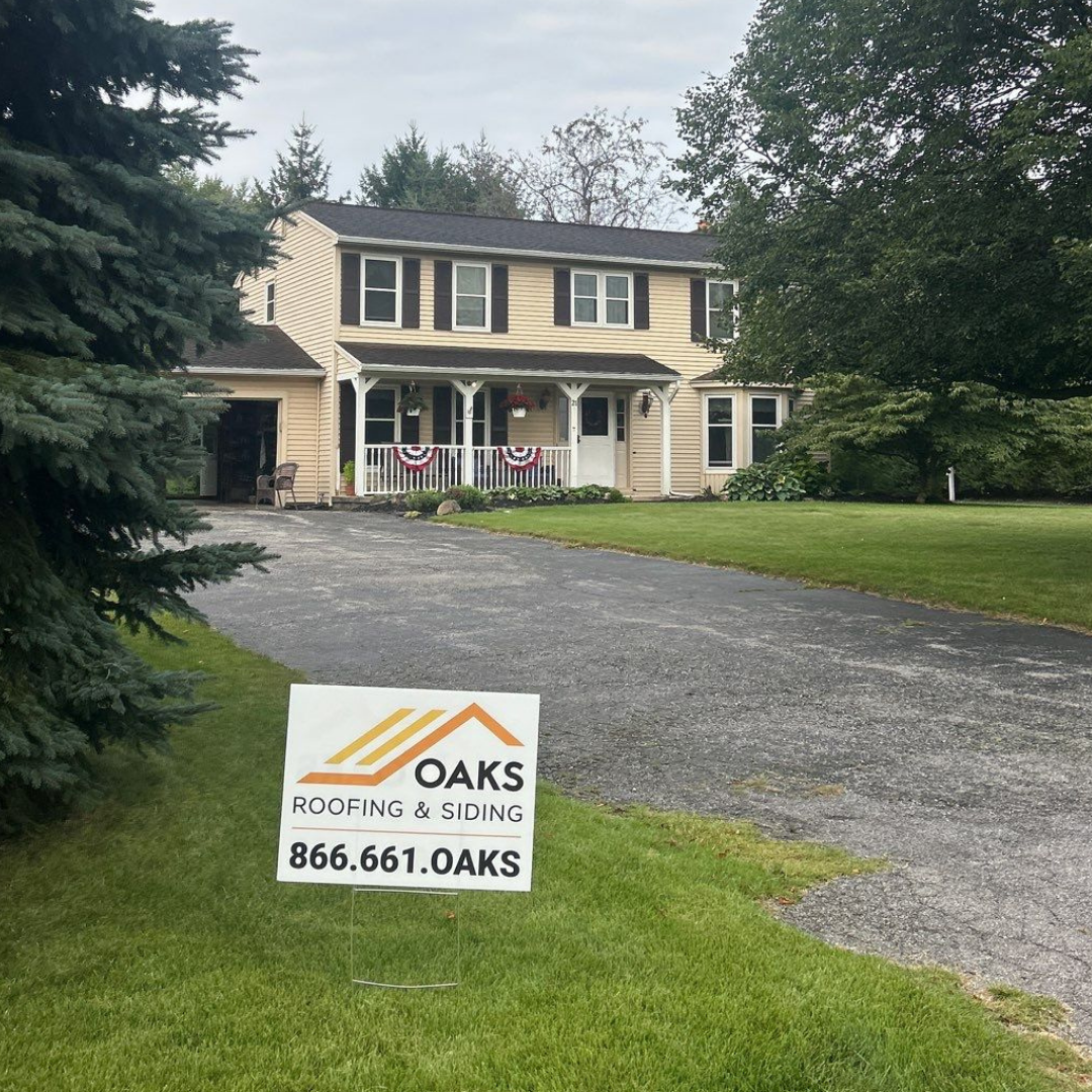 Oaks Roofing and Siding sign on a green lawn in front of a house with a newly installed roof.