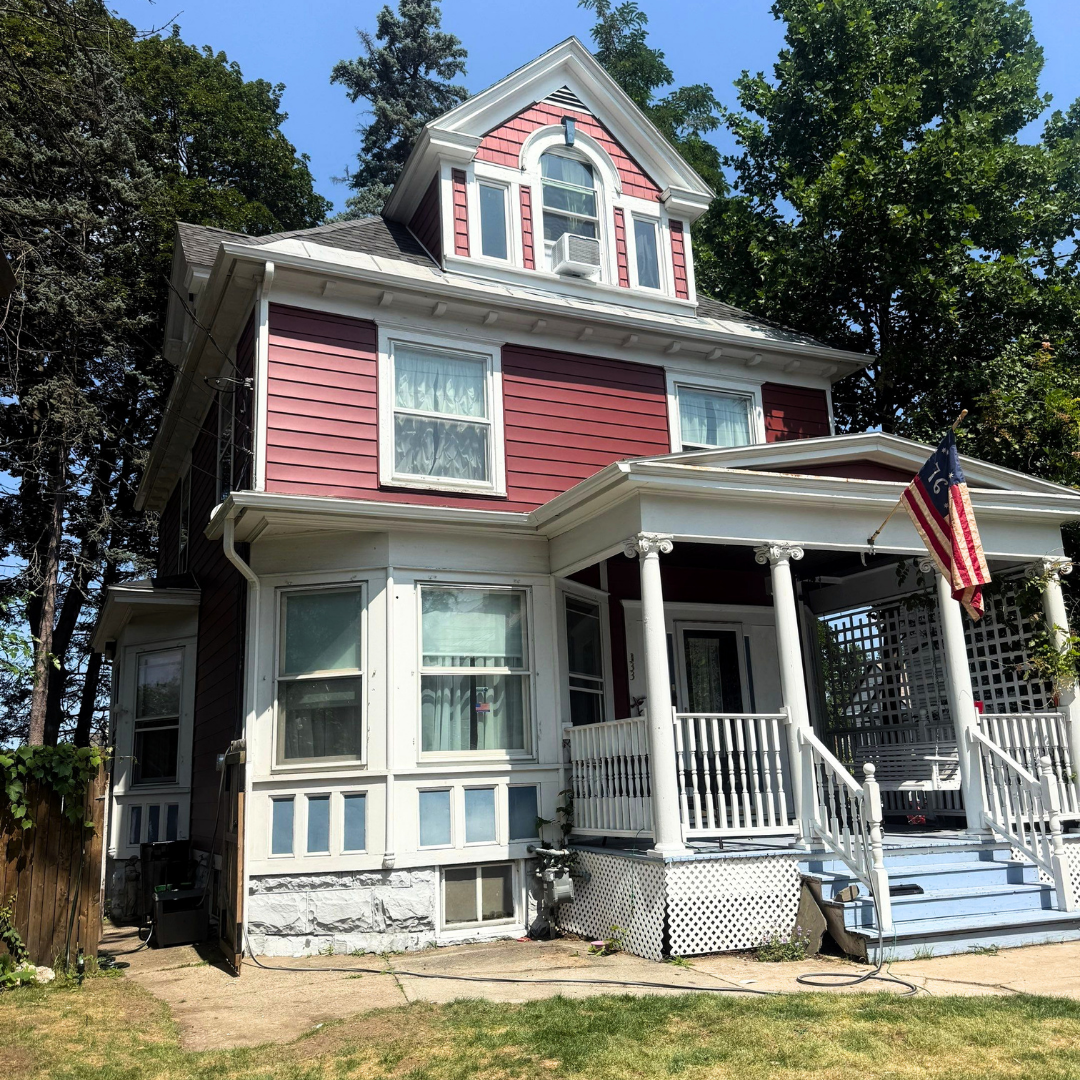 Exterior view of a two-story red house with white trim and a wrap-around porch. An American flag hangs near the entrance on a sunny day.