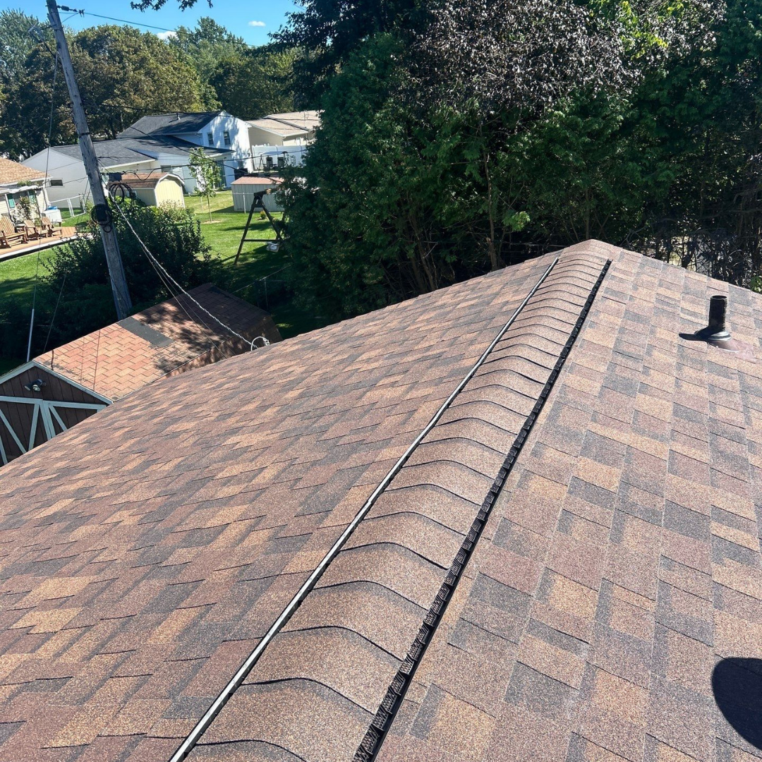 A close-up view of freshly installed brown asphalt shingles on a residential roof featuring a black ridge vent line running along the peak. The texture and pattern of the shingles are clearly visible against a backdrop of greenery.