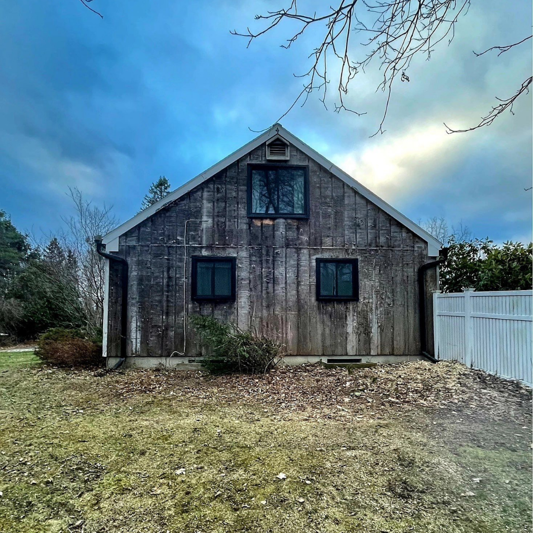 The image shows a rustic barn-style house with a gray, weathered wooden exterior under a dramatic blue sky. Bare trees and shrubs frame the property, which is bordered by a white vinyl fence.
