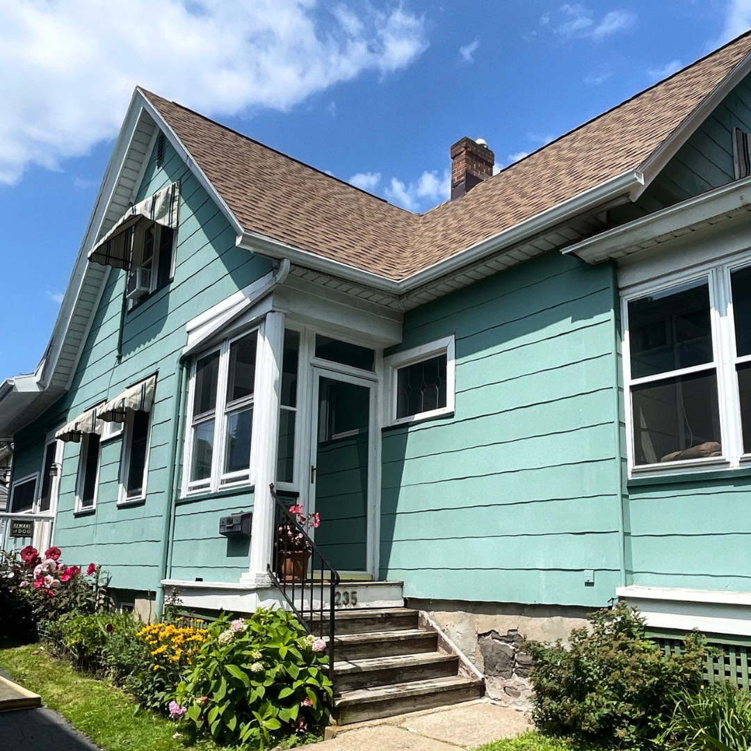 Beautiful teal Cape Cod home featuring a newly installed brown shingle roof. White trim, flower-filled garden, and bright blue sky complete the scene.