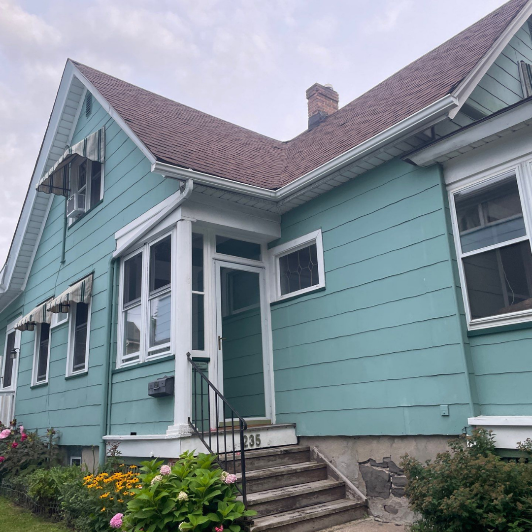 A quaint turquoise house with a steep roof and porch, surrounded by a colorful garden. The home features white trim, framed windows, and a stone foundation with a staircase leading to the entrance.