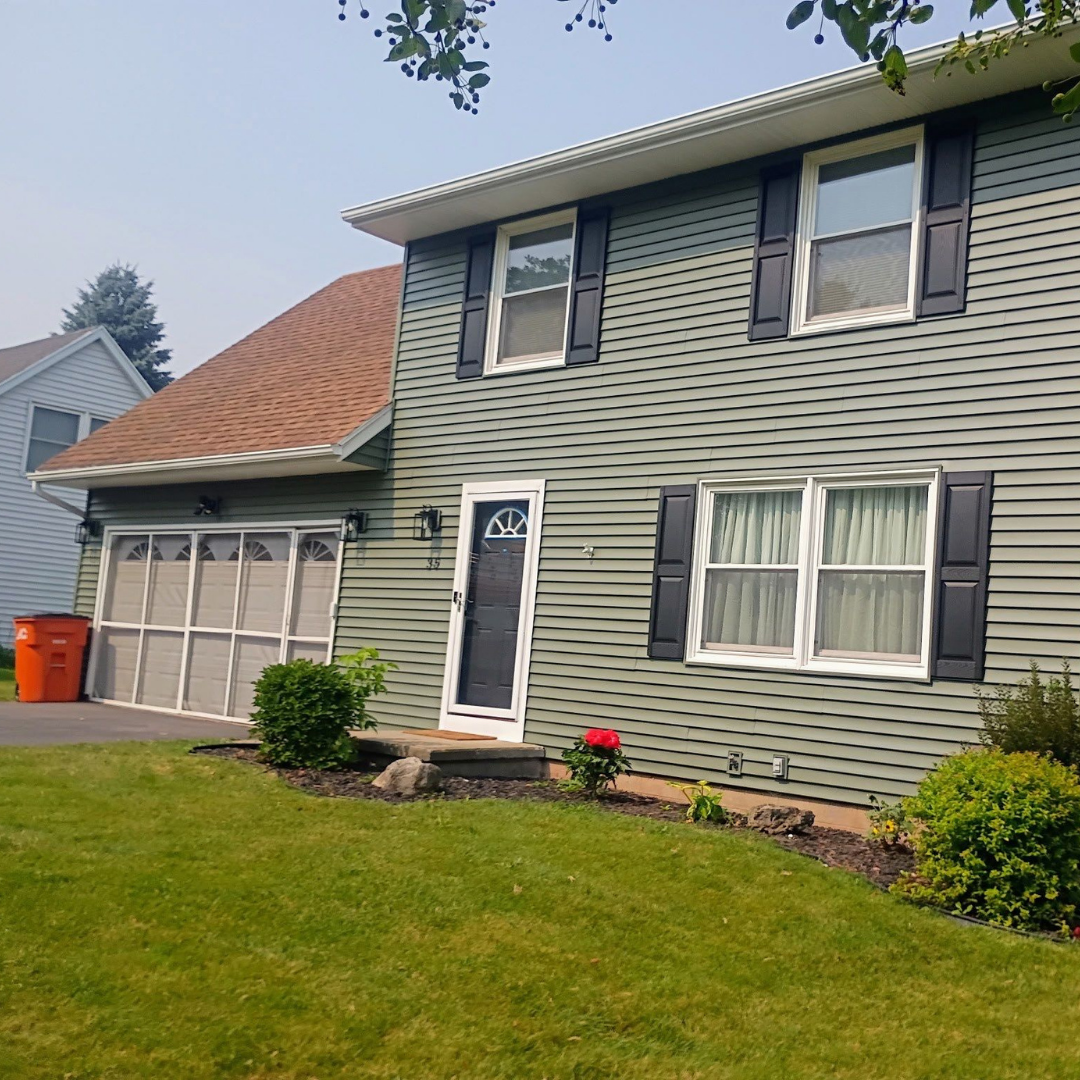 A two-story house with green siding and a brown roof, surrounded by a well-maintained lawn and small garden beds.