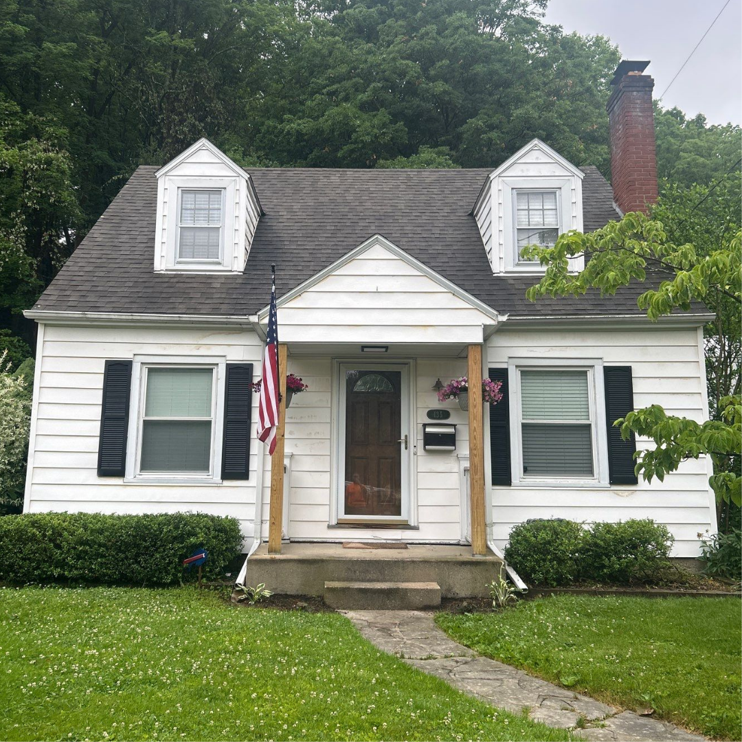 A charming white house featuring dormer windows and an American flag, set against lush greenery.